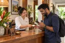 A receptionist gives a returning guest a blank return card as they check details on a phone.